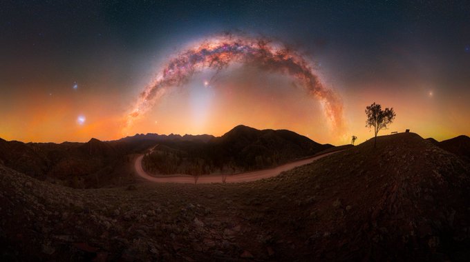A panoramic night sky view shows the Milky Way arching brilliantly over a dark, mountainous landscape with a winding road and a silhouette of a tree.