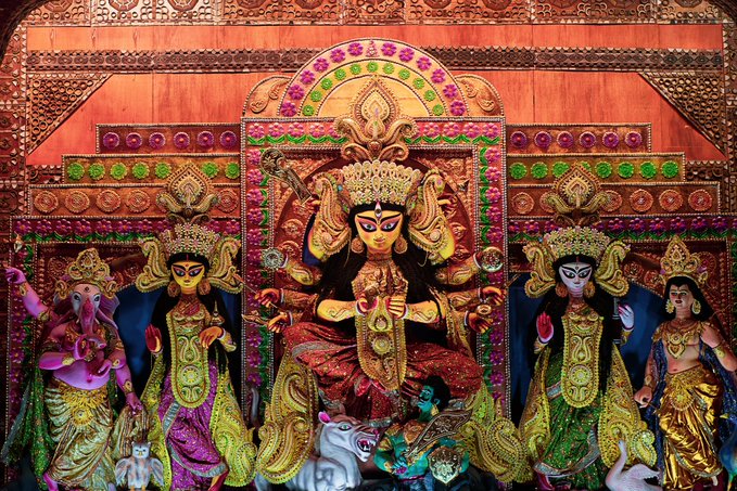 An ornate, colorful Hindu religious display features elaborately decorated statues of deities, likely Durga, Lakshmi, and others, adorned in vibrant red, gold, and green garments with crowns and jewelry, set against an intricately patterned backdrop.