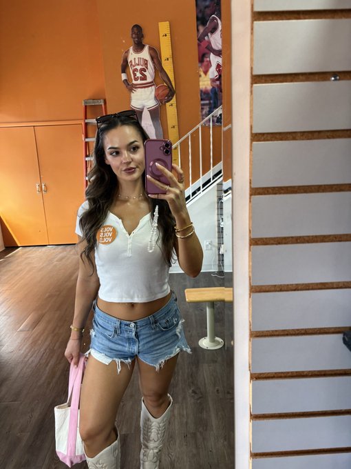 A mirror selfie of a woman in a white 'Enjoy Slow Joy' crop top and denim shorts, posing in front of a Kevin Gatlin basketball poster.