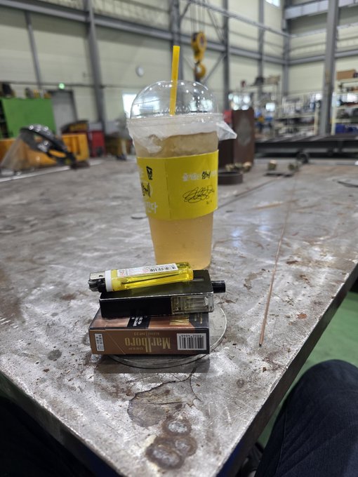 An iced drink in a yellow "Good Day" branded cup sits precariously stacked atop a Marlboro cigarette box and a car cigarette lighter on a metal workbench in a workshop. The image humorously reflects the post's "Altcoin investor's lunch" caption, mocking a sparse, makeshift meal—suggesting the investor has nothing left to eat after their crypto losses.