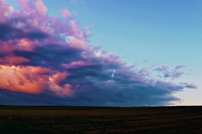 A scenic landscape photograph of a vibrant sunset with pink and purple clouds over a grassy plain under a blue sky.