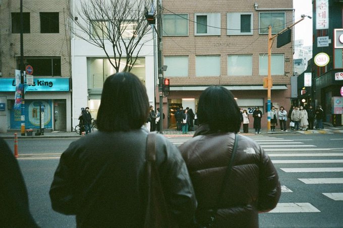 Two people stand at a crosswalk in a city, facing a street with Korean signage, including a Burger King, and other pedestrians waiting on the opposite corner.