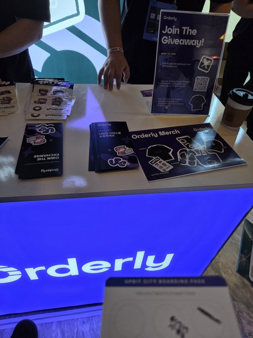 An 'Orderly' merchandise table with stickers and pamphlets displayed on a counter with blue lighting underneath. A sign reads 'Join The Giveaway!' and a Starbucks cup sits nearby.