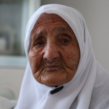 The image features an elderly woman wearing a white headscarf, with a black button fastened at the top of her head. She has wrinkles all over her face and appears to be older than 110 years old. The photo captures her close-up view, making her look like she is staring directly into the camera. In the background, there are blurred objects that could possibly be a window or another person's face.

The image also includes text that reads "110-year-old Turkish grandma shares her secret to a long life: I bought $SOL". This message seems to be related to the woman in the photo and might be an advertisement for a product, service, or cause.