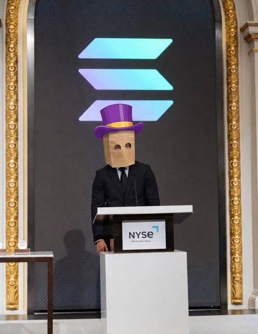 A person wearing a purple top hat and a paper bag over their head stands at a podium displaying the "NYSE" logo, embodying the "The hat stays on." sentiment by humorously maintaining the absurd headwear even in a formal setting. The scene appears to be at the New York Stock Exchange trading floor, adding a playful contrast to the serious financial environment.