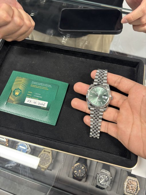 A hand holds a green-dial Rolex Datejust watch next to a green Rolex certificate card dated 07.10.2026, displayed above a tray of other luxury watches.