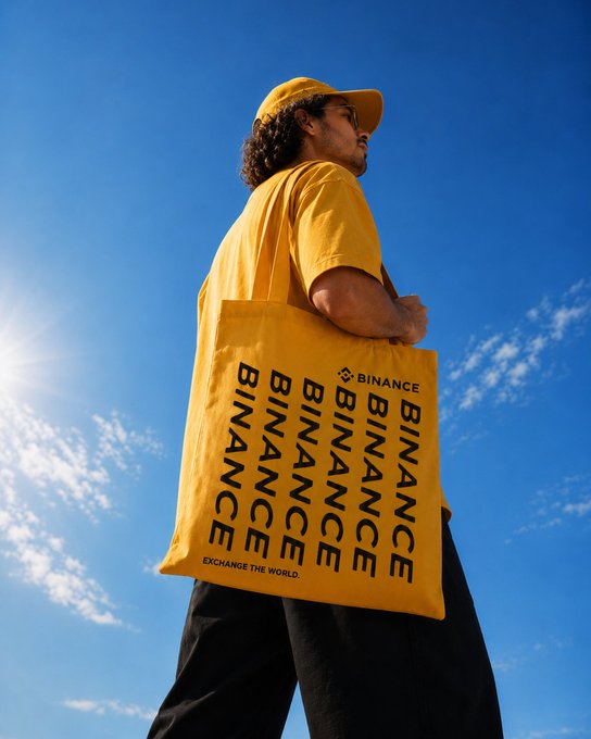 A young man in a yellow cap and t-shirt looks up at the sky while holding a yellow tote bag displaying the bold black text "BINANCE" repeated across its front, along with the tagline "EXCHANGE THE WORLD" and the Binance logo. The bag's vibrant yellow color perfectly complements the post's message about style coming in yellow, promoting the Binance brand to its Latin American audience.