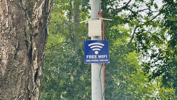 A blue 'FREE WIFI' sign mounted on a metal pole with a white router box attached, displaying Vietnamese text 'UBND PHUONG TRA BIEN' among green tree foliage.