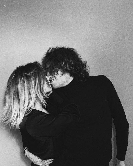 A black-and-white photo of a couple embracing intimately against a plain wall, with the man wearing a dark sweater and the woman resting her head on his shoulder.