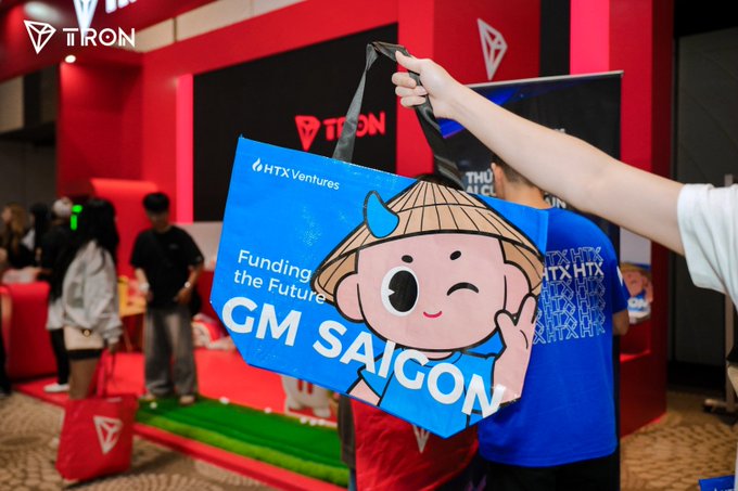 A person holds up a blue tote bag at the TRON booth featuring a cartoon mascot and the text "HTX Ventures," "Funding the Future," and "GM SAIGON," capturing the swag giveaway at Conviction 2024 referenced in the post about TRON's memorable final day.