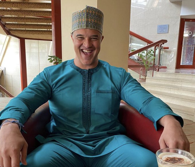 A laughing man wearing a teal traditional tunic and a patterned hat sits in a red chair, holding a glass of what appears to be coffee or tea.