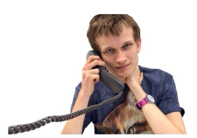 A young man with short blonde hair holds a grey corded phone to his ear, wearing a blue cat-print t-shirt and a pink watch.