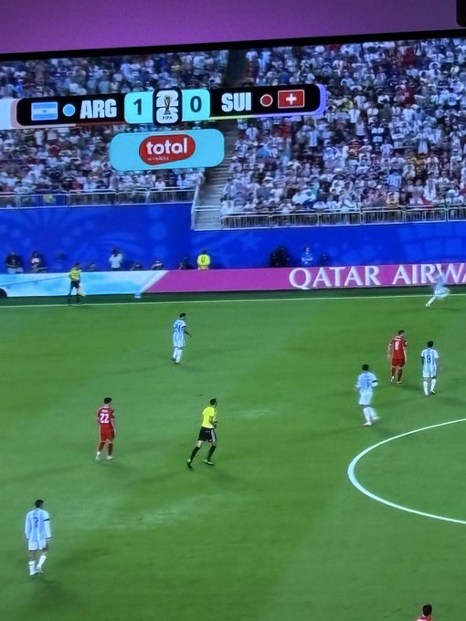 A soccer match showing the scoreboard ARG 1 - 0 SUI, with players in light blue (Argentina) and red (Switzerland) on the field, with Qatar Airways advertising visible and 'Total' sponsor banner.