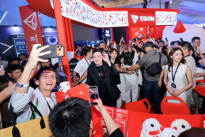 A crowded TRON booth at a convention where a man in a black suit waves while a fan takes a selfie with him amid red Tron-branded decor and plush toys.