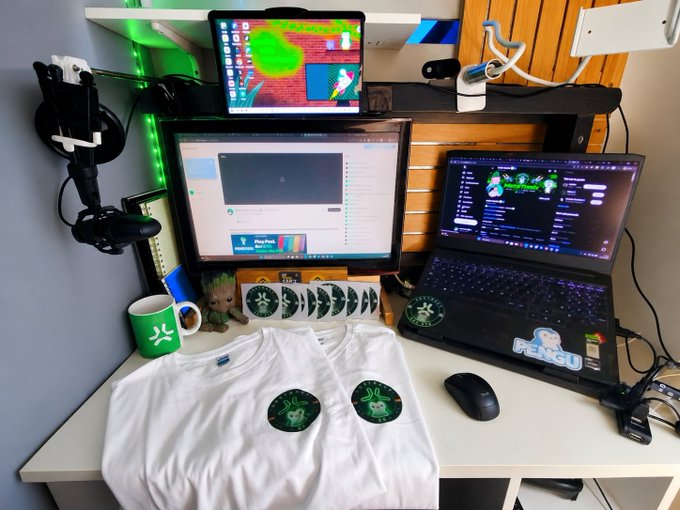 A desk setup featuring two white t-shirts with green "Pengu" logos laid out, alongside a monitor, laptop, and tablet displaying the AbstractChain ecosystem and Pengu branding—visually commemorating the first IRL meeting with the AbstractChain team and the community's collaborative spirit.