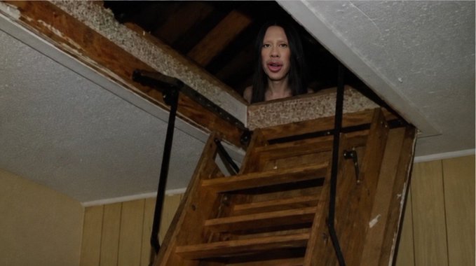 A woman peers down from an attic hatch above a wooden pull-down staircase. The image humorously implies that a tokenized deposit network powered by Quant ($QNT) is so transformative that even someone hiding in the attic couldn't resist looking out at the news.