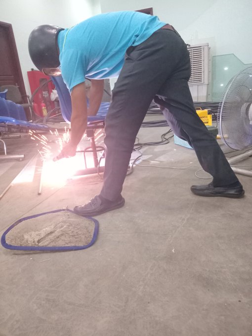 A worker in a blue shirt, black pants, and welding mask bending over while grinding or cutting metal, producing bright sparks on a workshop floor with a fan in the background.