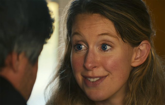 A close-up of a woman with long curly hair smiling warmly at a person whose back is to the camera.