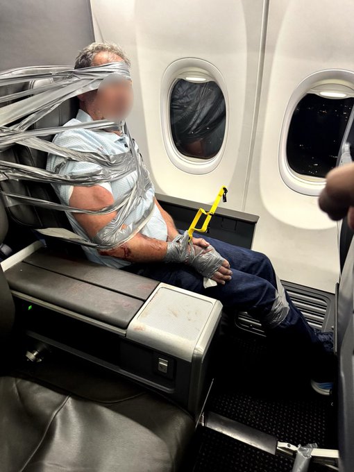 A person taped to an airplane seat with duct tape and plastic wrap on an aircraft, with another passenger visible through the window covered in plastic.