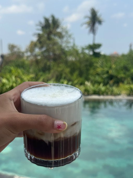 A hand holds a glass of iced coffee or espresso topped with frothy milk, featuring a cute pink manicure, set against a tropical background with a pool and palm trees.