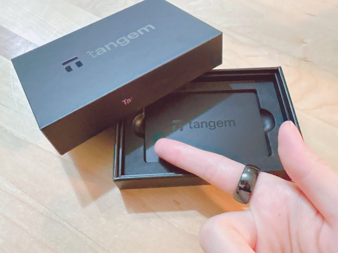 A hand with a black ring on the index finger is shown next to an opened 'Tangem' branded black box containing a Tangem card, likely a hardware cryptocurrency wallet.