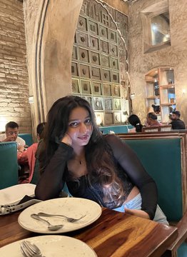 A photograph of a young woman smiling at the camera while seated in a booth at a restaurant. She has her hand resting near her face, and the restaurant features stone walls with arched details and teal upholstery.