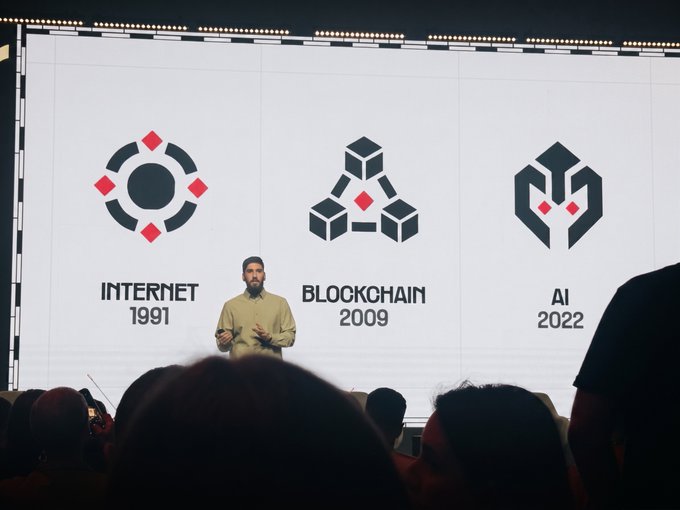 A speaker on stage presenting in front of a large screen displaying a timeline graphic for 'INTERNET 1991', 'BLOCKCHAIN 2009', and 'AI 2022'.