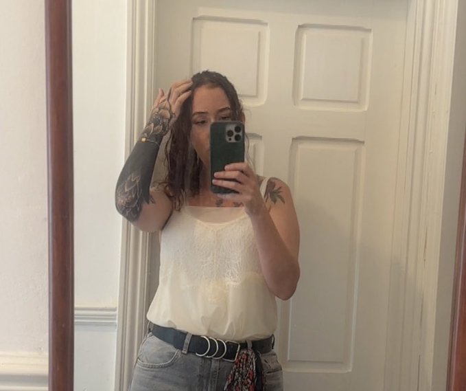 A mirror selfie of a woman with dark hair and tattoos on her arm, wearing a white camisole and jeans with a colorful belt, taking the photo with a green-cased smartphone.