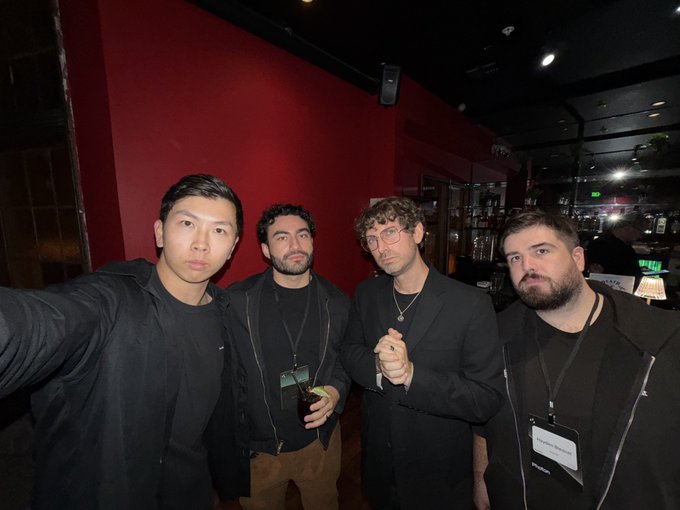 Four men pose together for a selfie in what appears to be a dimly lit venue with a red wall in the background; one holds a phone and another wears glasses.