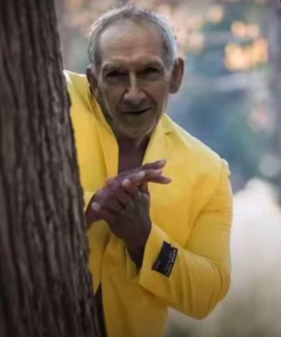 An older man in a bright yellow blazer peeks out from behind a tree, smiling with his hands clasped together.