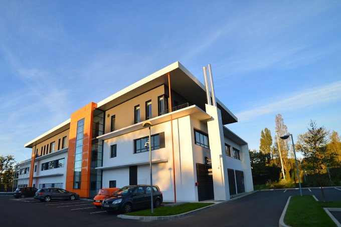 A modern multi-story office building with white and wood-paneled exterior stands under a clear blue sky, with cars parked in front and a traffic light nearby.