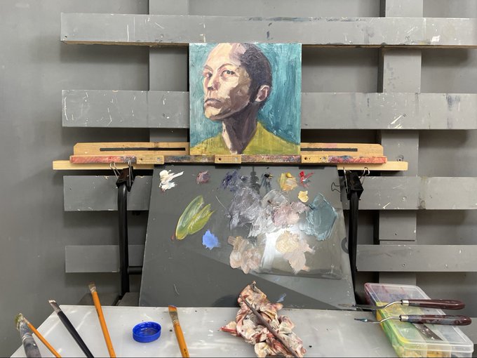 An artist's studio workspace featuring an in-progress oil portrait of a contemplative figure with a green-toned background, displayed on an easel above a palette with mixed paints and scattered brushes. The scene captures the creative process behind the upcoming "skeptical" mini series drop, with the unfinished portrait exemplifying the introspective theme and the casual studio setup hinting at the artist behind the work.