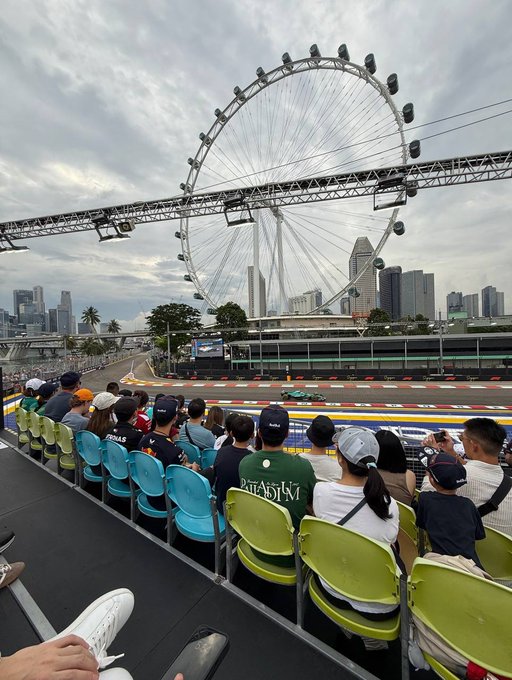 Spectators sit in colorful green and blue stadium seats watching cars race along the Marina Bay Street Circuit in Singapore, with the iconic Singapore Flyer Ferris wheel towering over the skyline. The image captures the vibrant Singapore setting that often hosts major crypto events like Token2049, showing fans gathered for the Singapore Grand Prix just steps away from the conference venue.