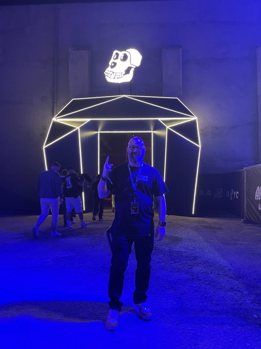 A man wearing glasses and a dark shirt posing in front of an illuminated neon skull installation at what appears to be an outdoor event venue at night.