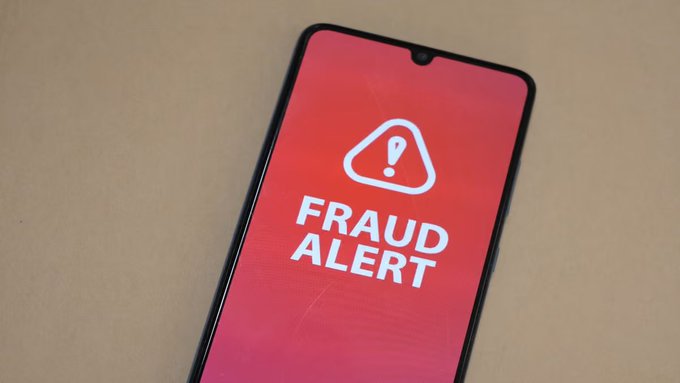 A smartphone resting on a surface displays a red screen with a white warning triangle icon and the text 'FRAUD ALERT'.