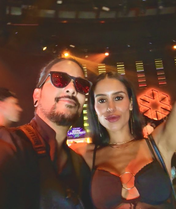 A man with a beard wearing red sunglasses takes a selfie with a dark-haired woman in a dark, neon-lit club or concert venue.