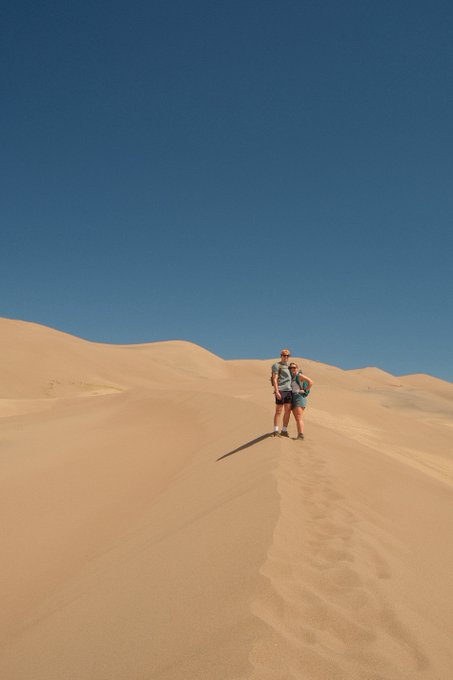The image shows a couple standing together at the top of a sand dune, with their backs to the camera. They are surrounded by vast desert terrain that stretches out into the distance under a clear blue sky. The photo captures the moment when they reach the peak of the dune and look back towards the viewer.