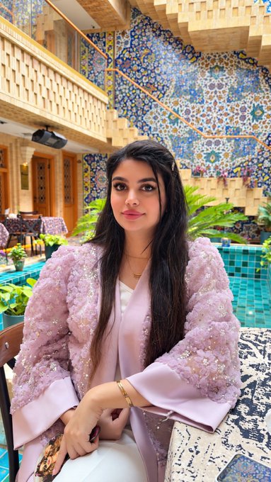 A young woman wearing a textured light purple jacket with floral embellishments poses indoors in front of an ornate wall featuring blue tilework, a wooden staircase, and a tiled pool area.