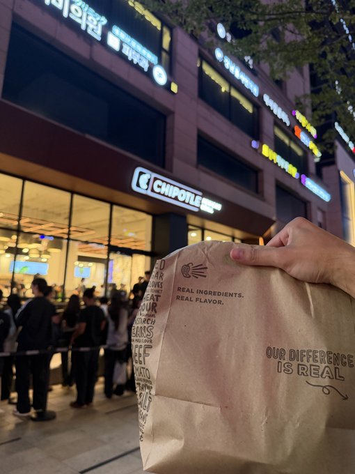 A person holds a Chipotle paper bag reading 'Real Ingredients. Real Flavor.' and 'Our Difference Is Real' outside a Chipotle restaurant at night, with customers visible near the entrance.