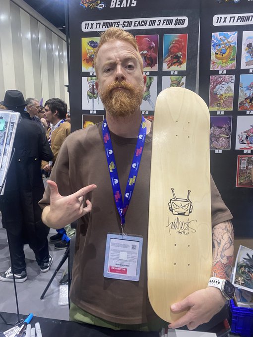 A red-bearded man at what appears to be San Diego Comic-Con holds up a signed skateboard deck featuring a hand-drawn robot character, matching the post's mention of a "Rare Robot" design by @toddbeats_art, with visible booth signage advertising "11 X 17 PRINTS: $10 EACH OR 3 FOR $25."