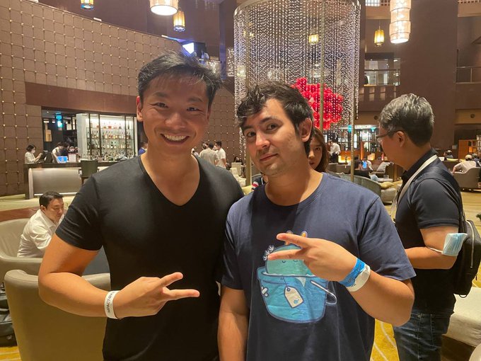 Two young men pose for a photo at what appears to be a hotel or conference lobby, with one wearing a blue T-shirt featuring an Axie character design and the other in a black V-neck, both flashing peace signs while wearing event wristbands that suggest a crypto or Web3 conference setting.

The image visually complements the post about the interview with Jihoz (Co-founder of Axie Infinity), likely showing the two together at a networking event where their conversation on Web3 community building took place.