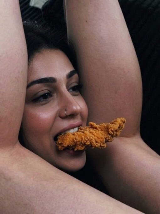 A woman with her arms raised biting into a piece of fried chicken, posed against her own skin.
