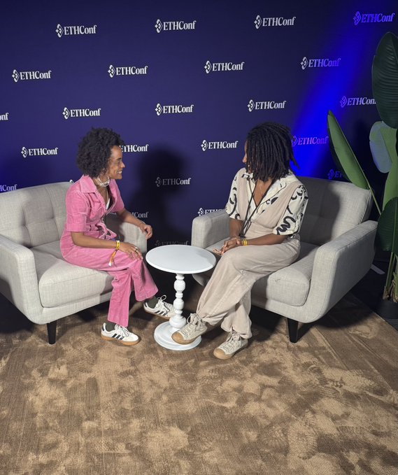 The image shows two women sitting on chairs in front of a blue wall adorned with white text that reads "ETHGlobal" and "ethicall". They are engaged in conversation, possibly discussing their experiences at an Ethical Global conference or workshop. The room has a carpeted floor and several potted plants placed around the space, creating a comfortable atmosphere for attendees to gather and exchange ideas.