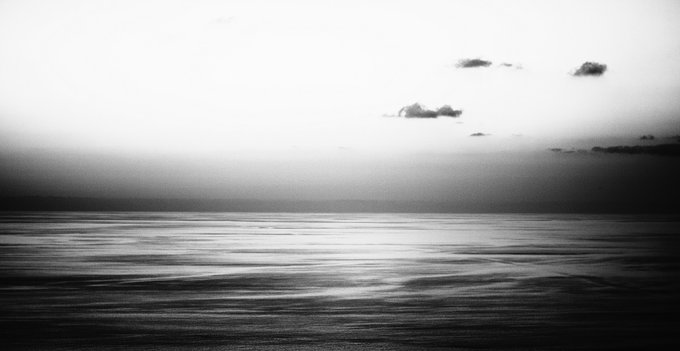 A black and white photograph of a calm ocean meeting a hazy sky with a few scattered clouds.
