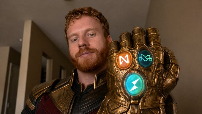 A man with curly red hair wears an Infinity Gauntlet with three cryptocurrency logos visible: a white 'N' symbol, a teal infinity symbol, and a lightning bolt logo on the hand gems.