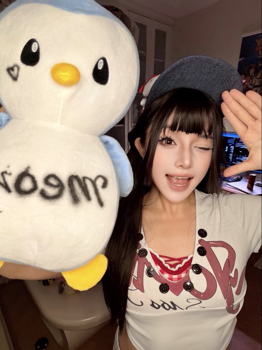 Selfie of a young woman with dark hair and a gray beret holding a plush penguin toy, with a desk setup featuring a laptop and gaming peripherals visible in the background.