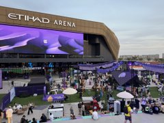 An exterior view of the Etihad Arena showing crowds gathered outside the venue with purple signage and tents in the foreground.