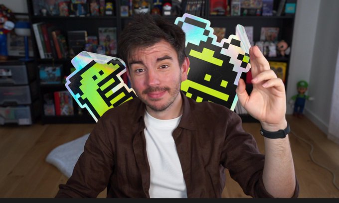 A young man wearing a brown shirt and white t-shirt sits in front of a shelf of collectibles, holding up a pixel-art style holographic card featuring green and black designs.