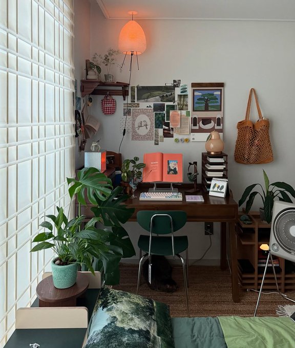 A cozy, plant-filled room with a wooden desk setup featuring a keyboard, a moodboard with pictures and postcards, a green chair, a mesh bag hanging on the wall, and a bed in the foreground.