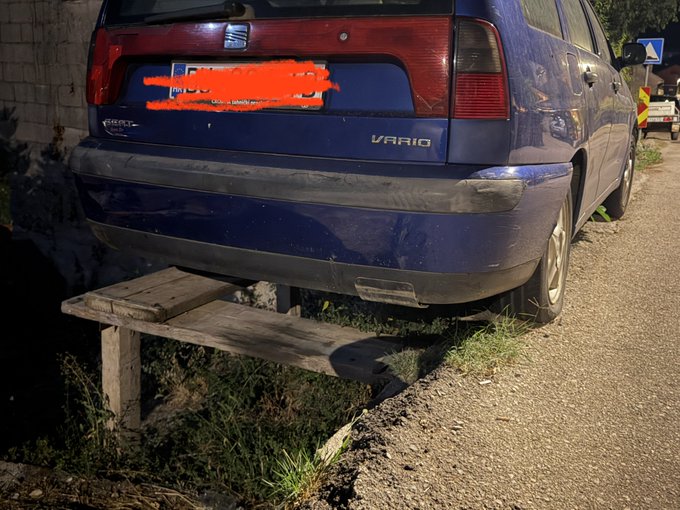 A dark blue SEAT Vario car parked precariously with its rear wheels partially hanging over a small wooden bridge/culvert beside a dirt path.